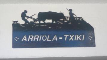 Silueta de una persona con un perro y una vaca en un vehículo, y el texto 'ARRIOLA-TXIKI'.