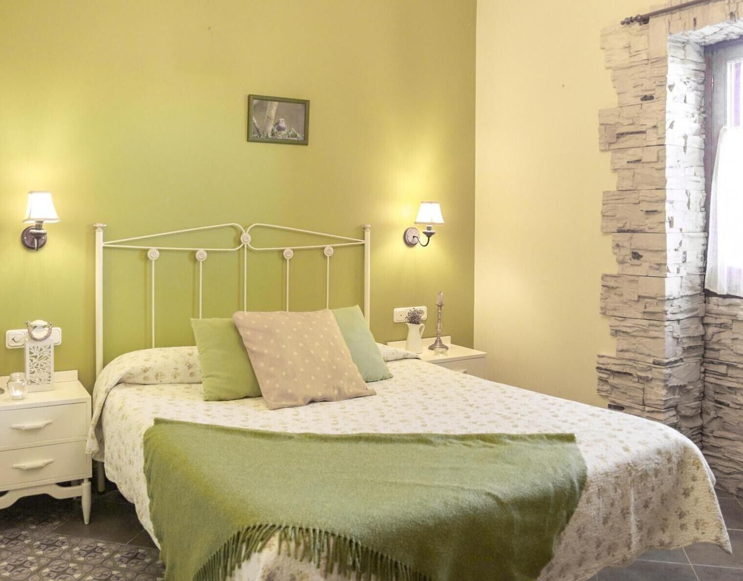 A bed with a metal frame, two bedside tables, a wall-mounted lamp, a small picture, and a stone wall.