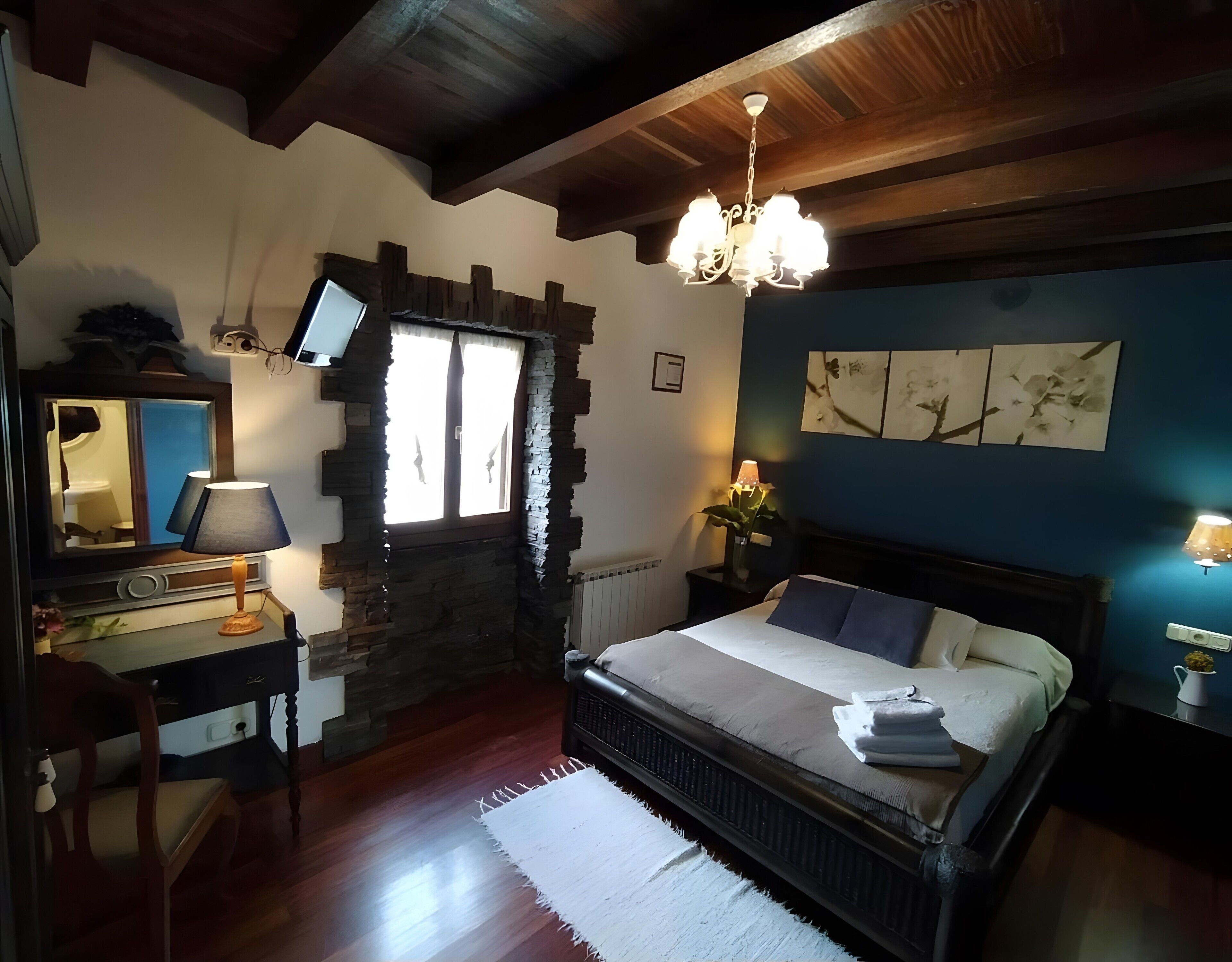 Ein Schlafzimmer mit Steinwand, Holzboden, einem Bett mit Kissen, einem Nachttisch mit Lampe, einer Kommode und einem Spiegel.