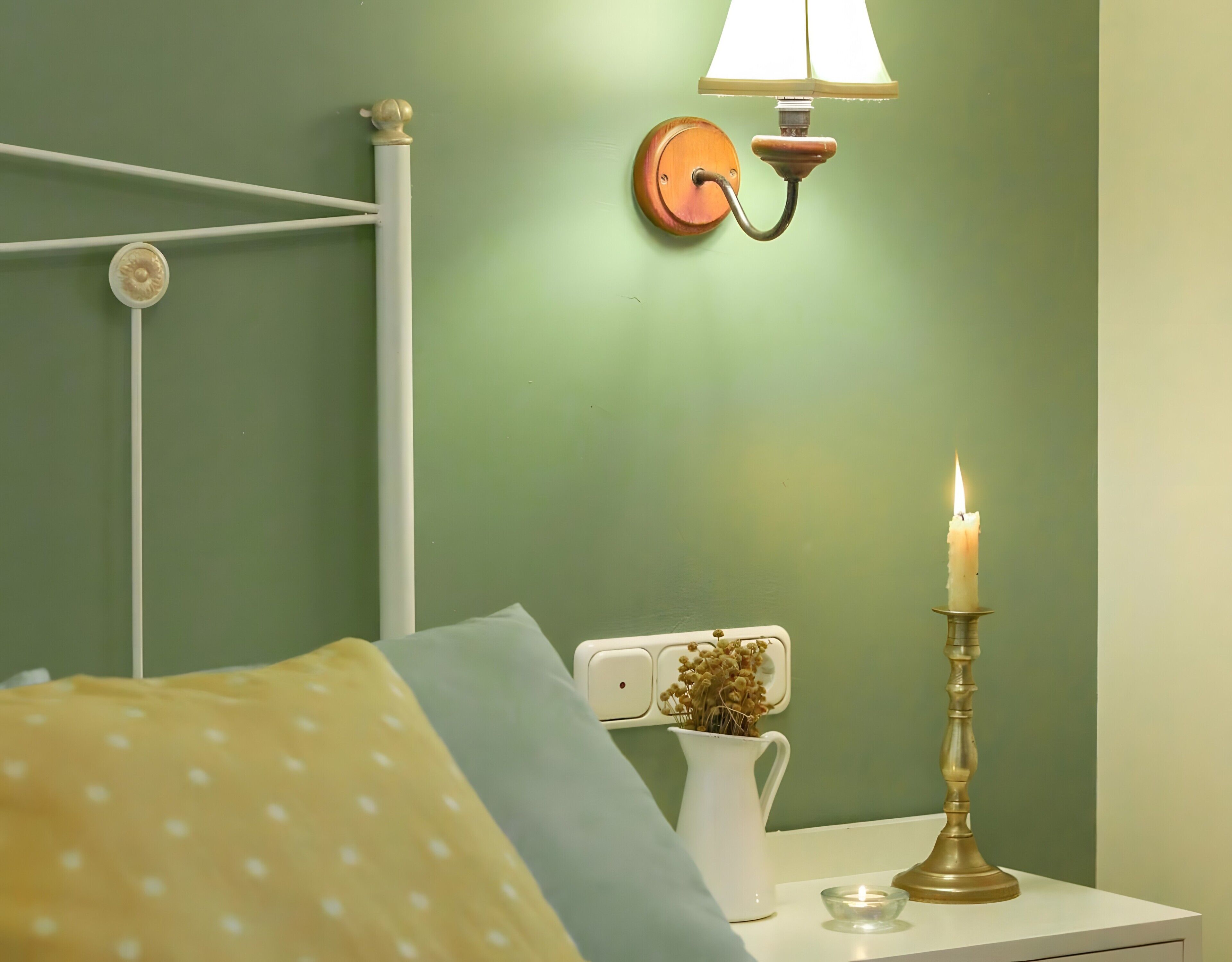 Een slaapkamer met een bed, een nachtkastje met een lamp, een wandlamp, een kaars en een vaas met gedroogde bloemen.