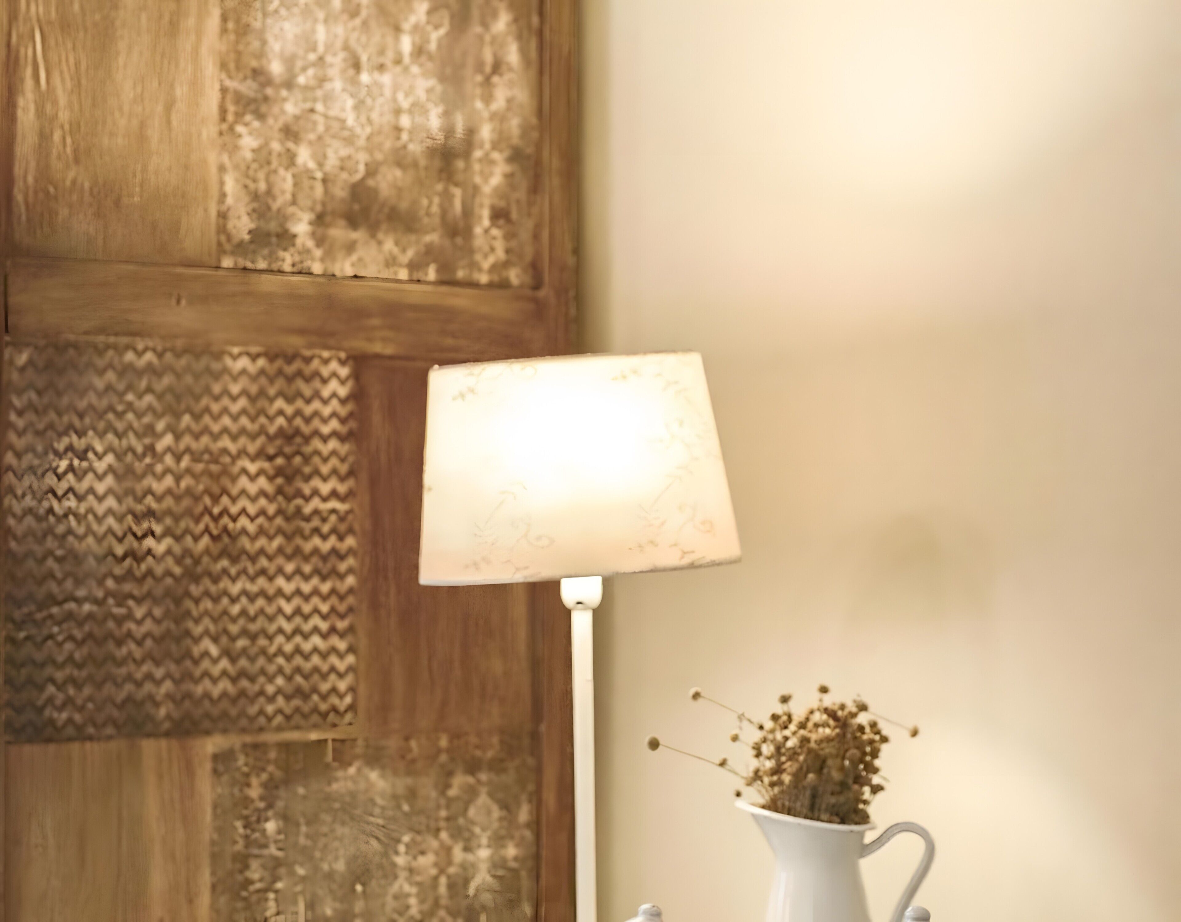 A lamp with a white shade, a white vase with dried flowers, and a wooden door with a textured panel.