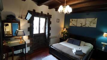 Un dormitorio con pared de piedra, techo de madera, una cama con almohadas, una mesita de noche con lámpara, un aparador y un espejo.