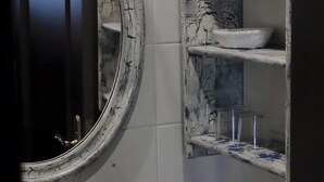 Ein Badezimmer mit weiß geflieste Wand, einem weißen Waschbecken mit Marmor-ähnlicher Rückwand und einer weißen Vase mit getrockneten Blumen.