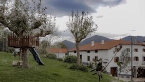Una casa arbre amb tobogan, zona enjardinada i edificis amb teules vermelles.