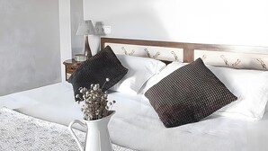 Un llit ben fet amb dos coixins decoratius, una làmpada de nit i un ram de flors seques sobre una catifa blanca amb textura.
