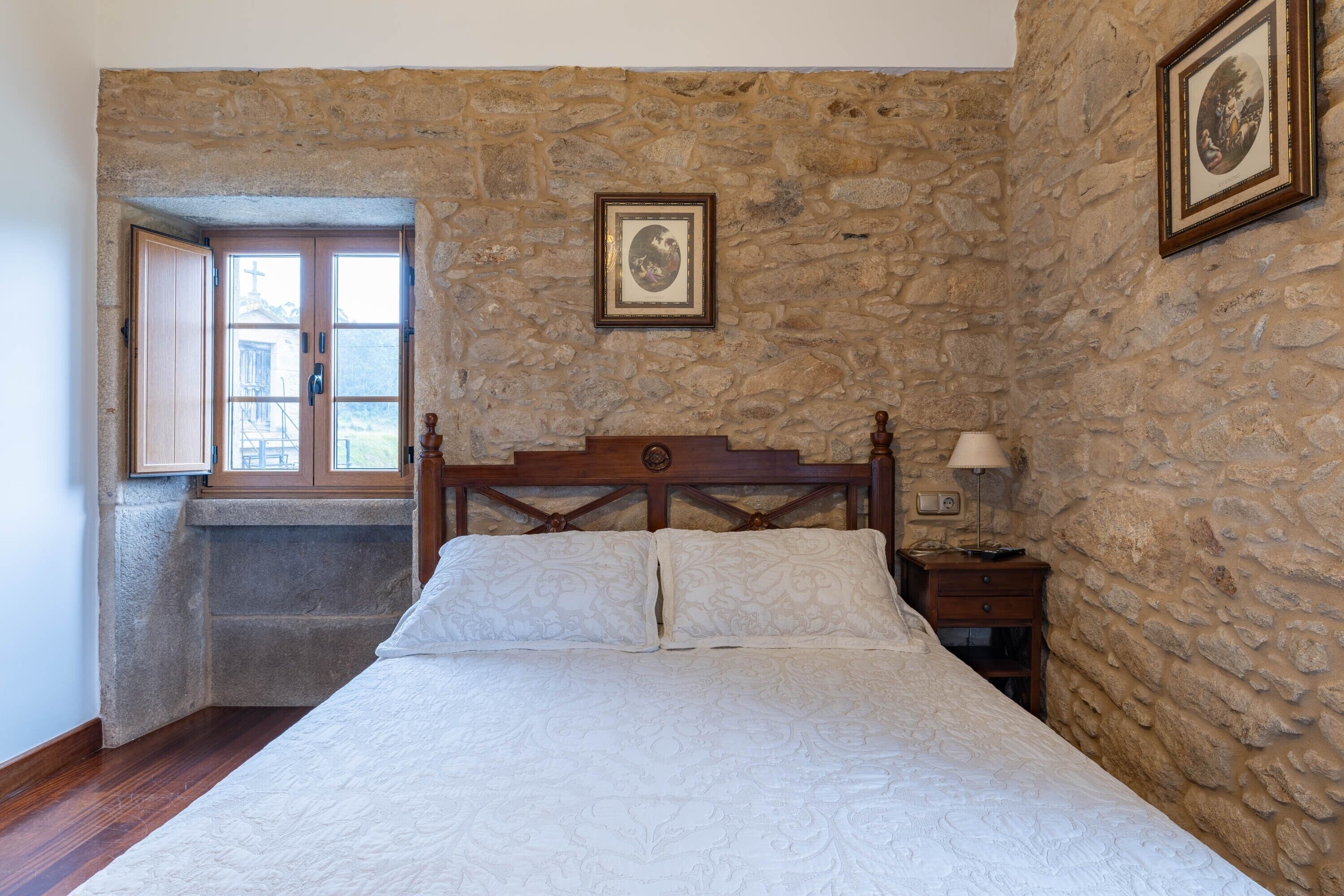 A bedroom with a stone wall, a wooden bed frame, a nightstand with a lamp, and a window with a view of a building outside.