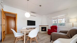 Een woonkamer met een open haard, een aan de muur bevestigde televisie, een eettafel met stoelen en een rode fauteuil.