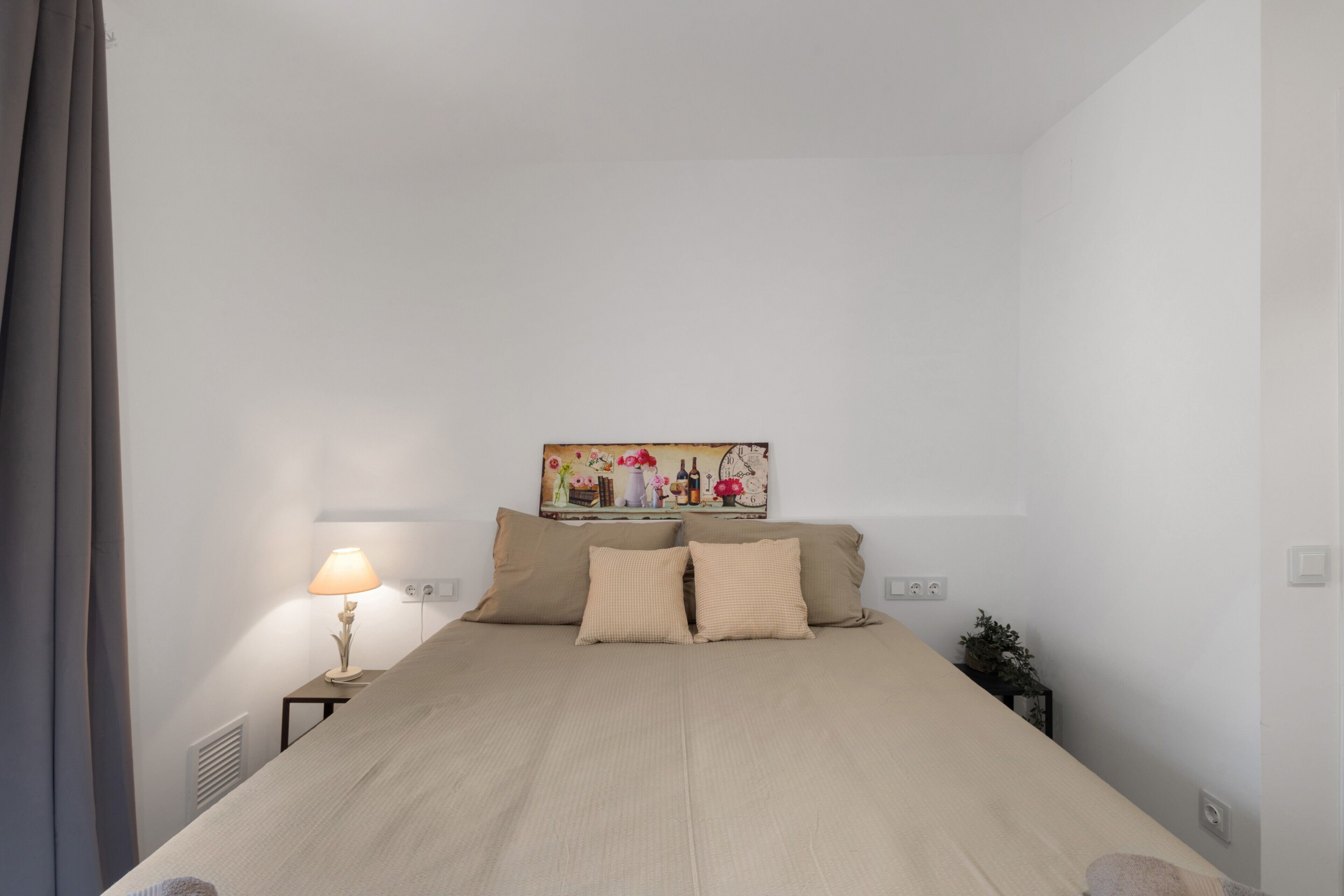 A bedroom with a bed, a nightstand with a lamp, a wall-mounted painting, and a potted plant.
