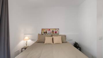 A bedroom with a bed, a nightstand with a lamp, a wall-mounted painting, and a potted plant.