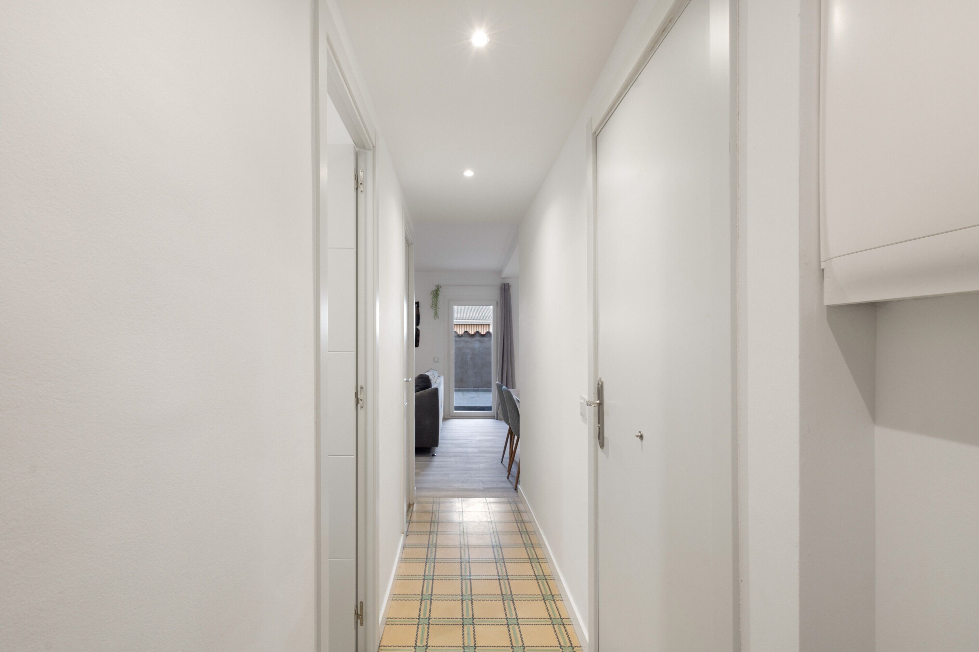 Un pasillo estrecho con paredes blancas, piso a cuadros y una puerta que da a una habitación con cama y ventana.