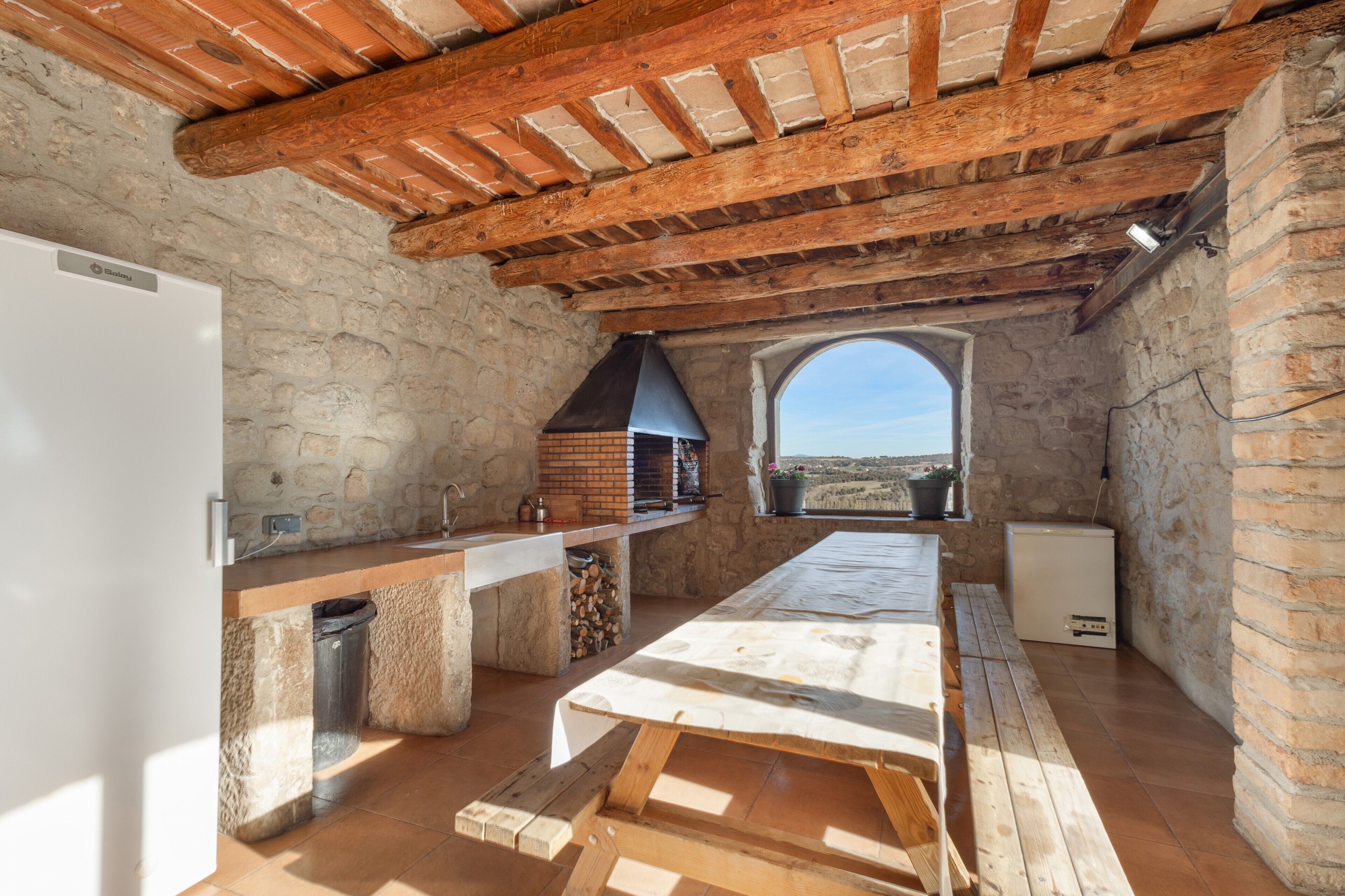 A rustic kitchen with a stone wall, wooden beams, a brick oven, and a view through an archway.