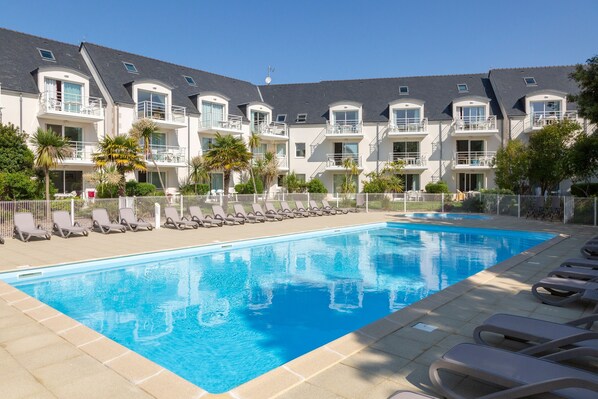 A swimming pool with lounge chairs, a building with balconies, and a clear sky.
