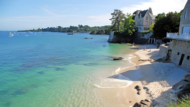 Una spiaggia costiera con acqua cristallina, una riva sabbiosa e un grande edificio con un balcone che si affaccia sul mare.