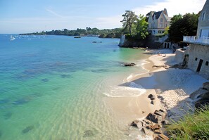 A coastal beach with clear water, a sandy shore, and a large building with a balcony overlooking the sea.