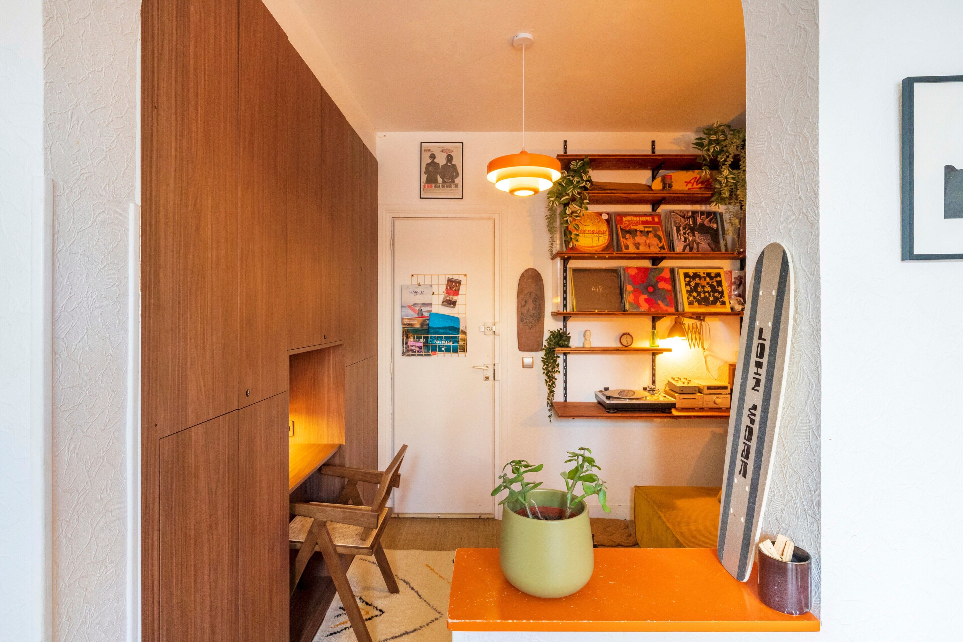 Une pièce avec une armoire en bois, une petite table sur laquelle est posée une plante, un skateboard et un mur orné de tableaux et sur lequel est fixée une lampe.