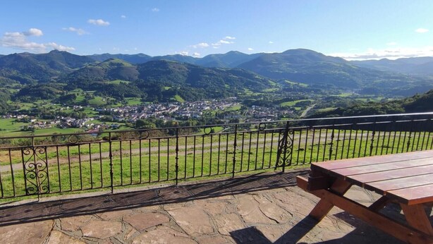 A scenic view from a balcony with a wooden table, overlooking a town nestled in green valleys and mountains.