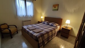 A bedroom with a bed, two bedside tables with lamps, a chair, a radiator, and a window with curtains.