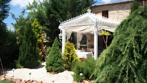 A gazebo with a white canopy, surrounded by evergreen trees and a stone building in the background.