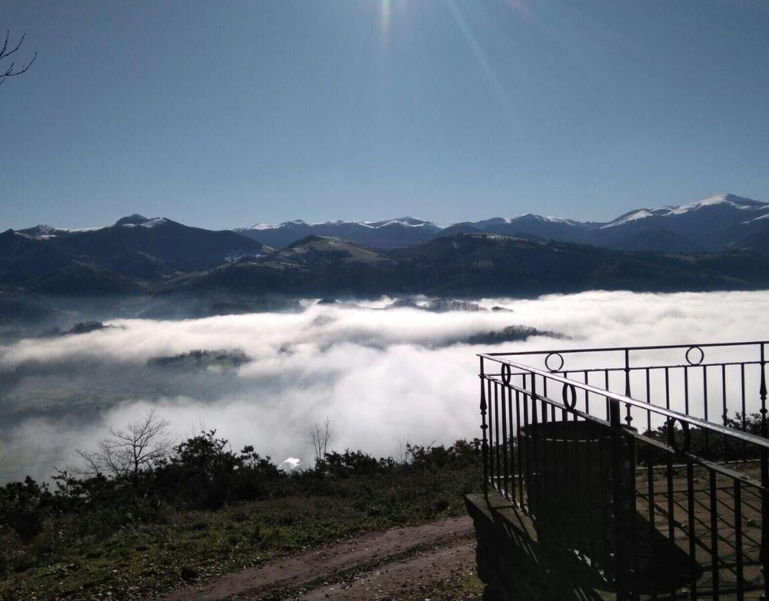A mountain landscape with a clear sky, a railing, and a path leading towards the mountains.