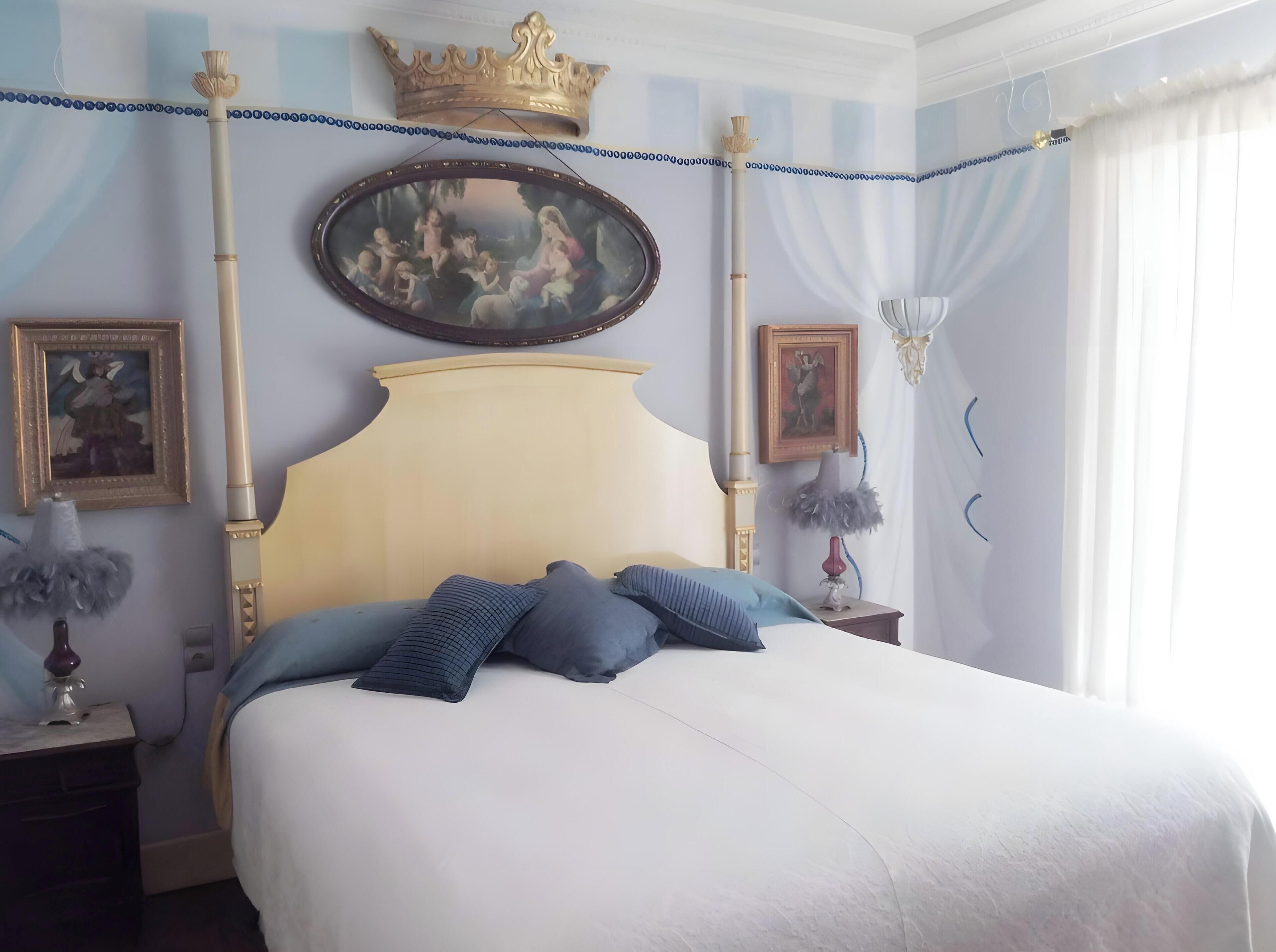 A bedroom with a bed, a headboard, two bedside tables, a wall-mounted crown, a framed painting, and a window with curtains.