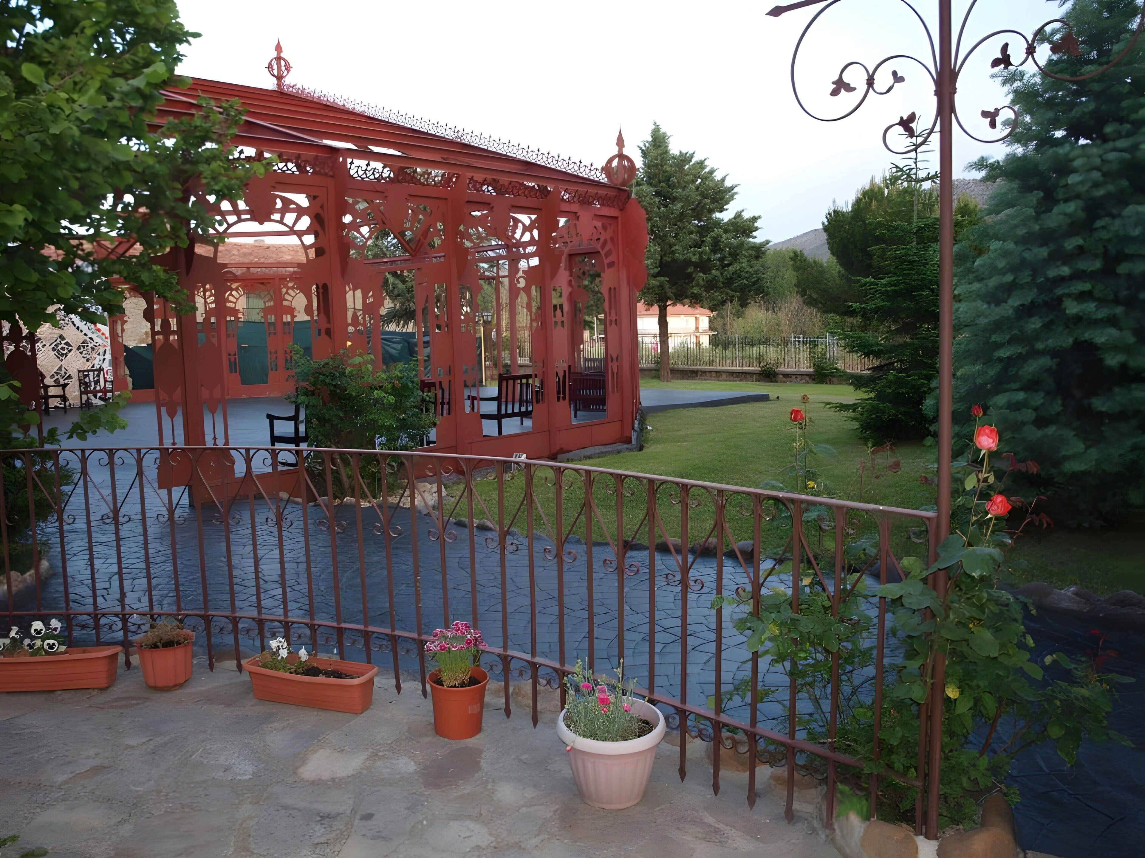 Un mirador rojo con techo estampado, rodeado por una valla de metal y macetas con plantas en primer plano.