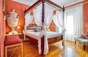 Un lit à baldaquin à quatre poteaux, agrémenté de rideaux blancs, un grand vase, un banc rouge et un canapé, le tout dans une pièce au plancher en bois et aux murs orange.