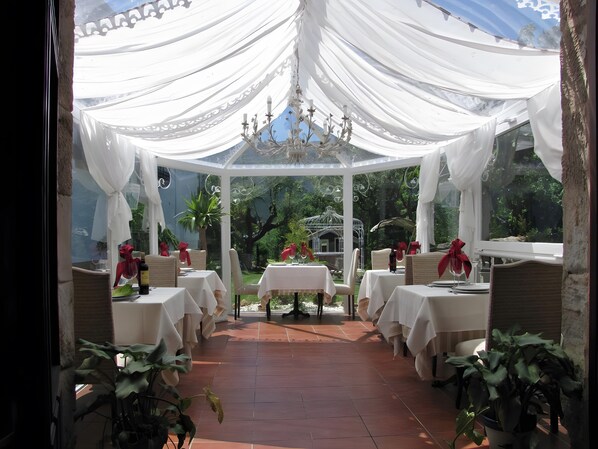 A covered outdoor dining area with tables set for two, surrounded by potted plants.