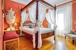 Una cama con dosel adornada con cortinas blancas, un jarrón grande, un banco rojo y piso de madera.
