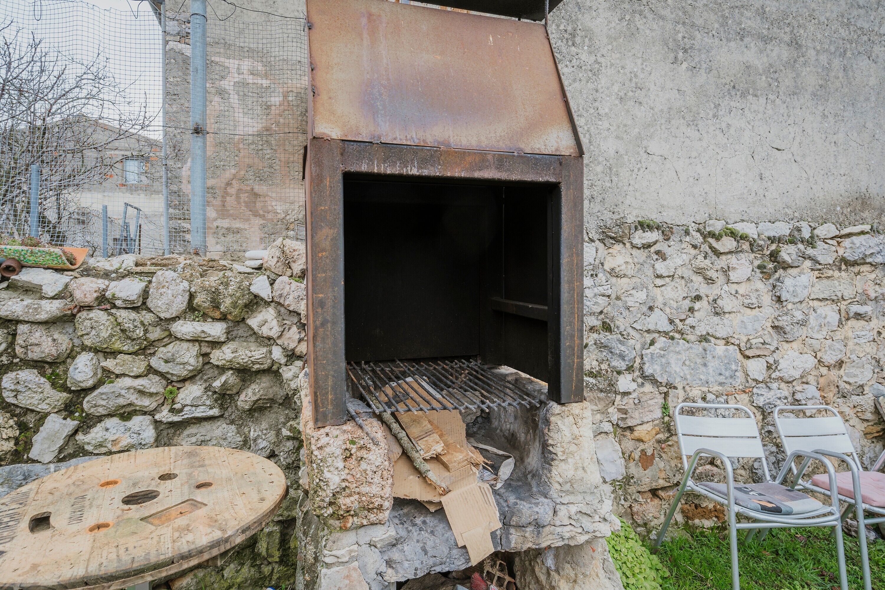 Un barbecue rustique installé à l’extérieur, avec un mur de pierres en arrière-plan, une chaise en métal et une table ronde.