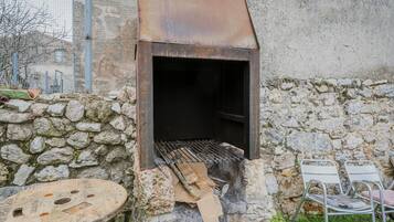 Un barbecue rustique installé à l’extérieur, avec un mur de pierres en arrière-plan, une chaise en métal et une table ronde.