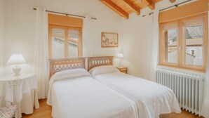 Une chambre avec deux lits, un plafond en bois, une fenêtre avec des volets, une table de chevet avec une lampe et un radiateur.