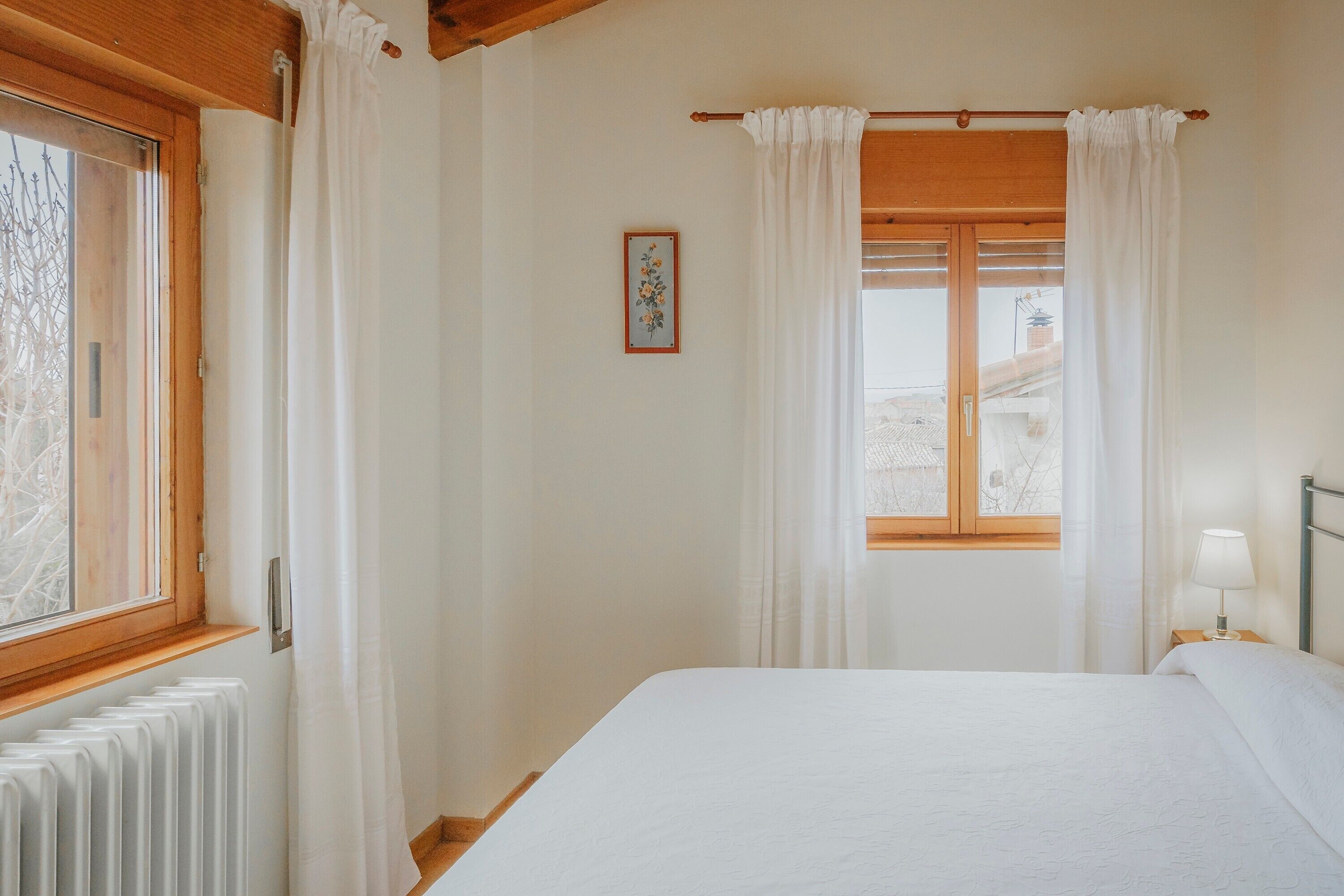 Quarto com cama, janela com cortinas, radiador e mesa de cabeceira com abajur.