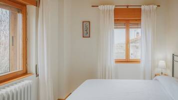 Quarto com cama, janela com cortinas, radiador e mesa de cabeceira com abajur.
