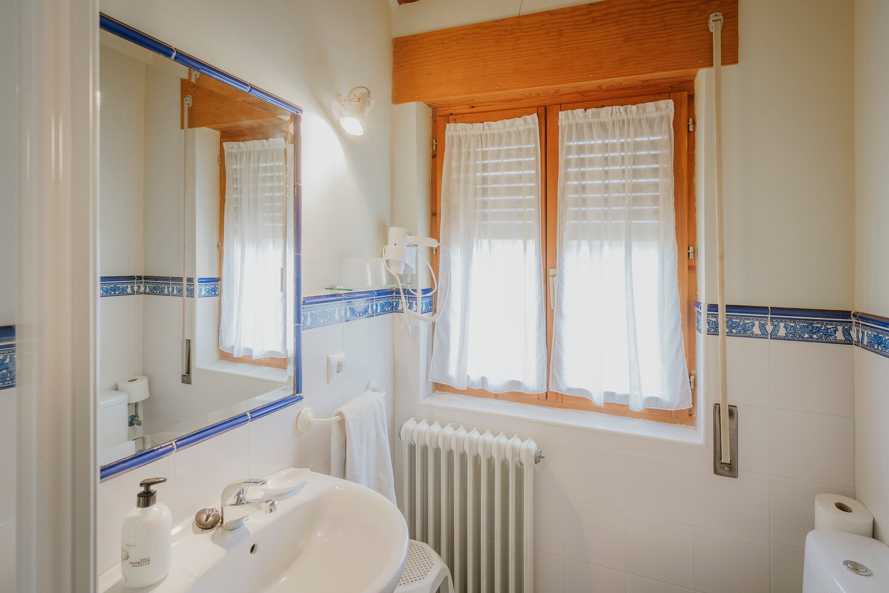 Un baño con un espejo grande, un lavabo, un inodoro, una ventana con cortinas y un radiador.
