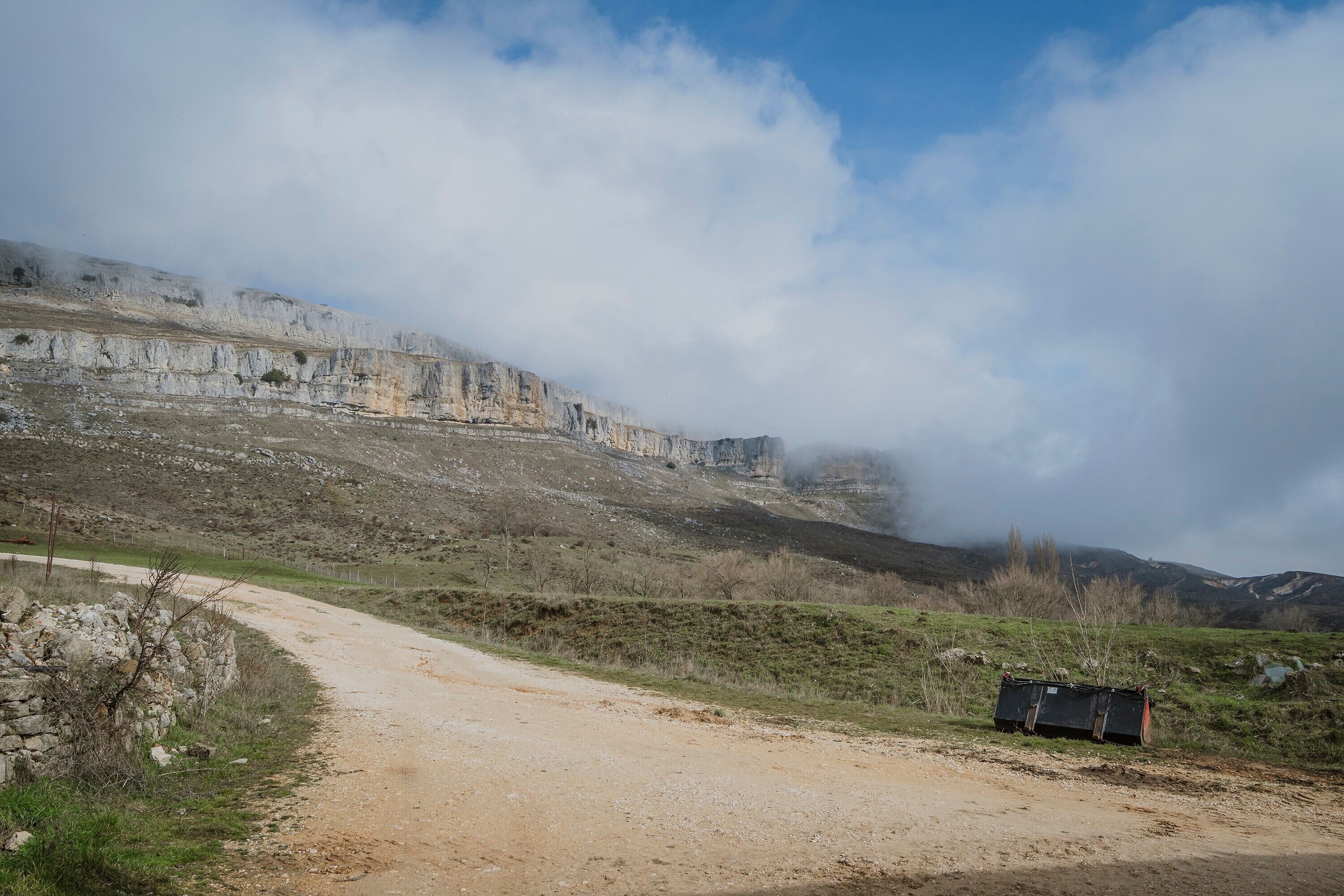 A dirt road leading to a cliff with a stone wall on the left and a metal structure on the right.