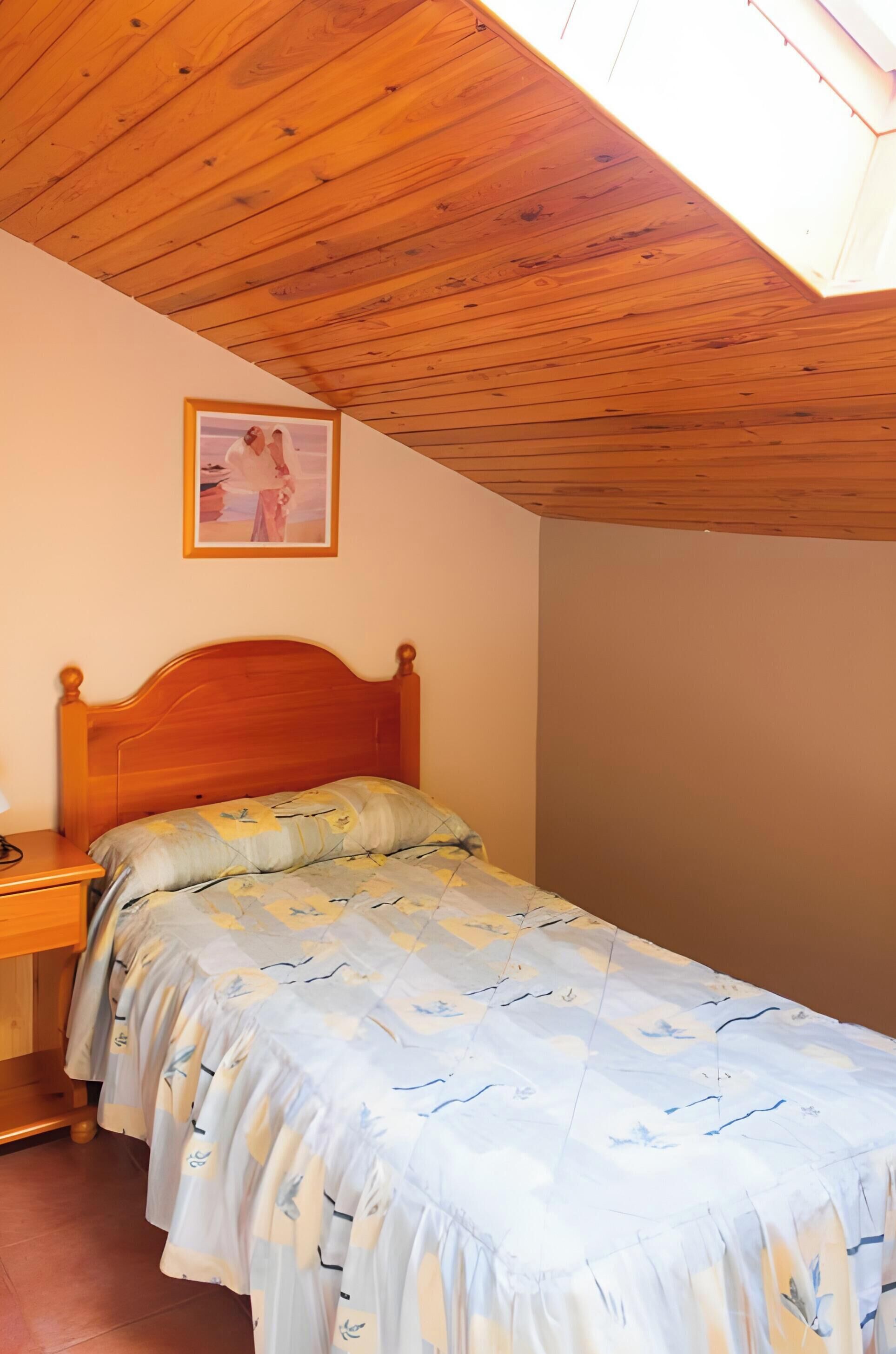 Un dormitorio con techo de madera, una cama con colcha floral, una mesita de noche y un cuadro enmarcado en la pared.