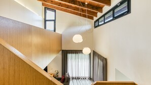 A modern interior with a wooden staircase, a seating area with a sofa and curtains, and a ceiling with exposed beams and pendant lights.