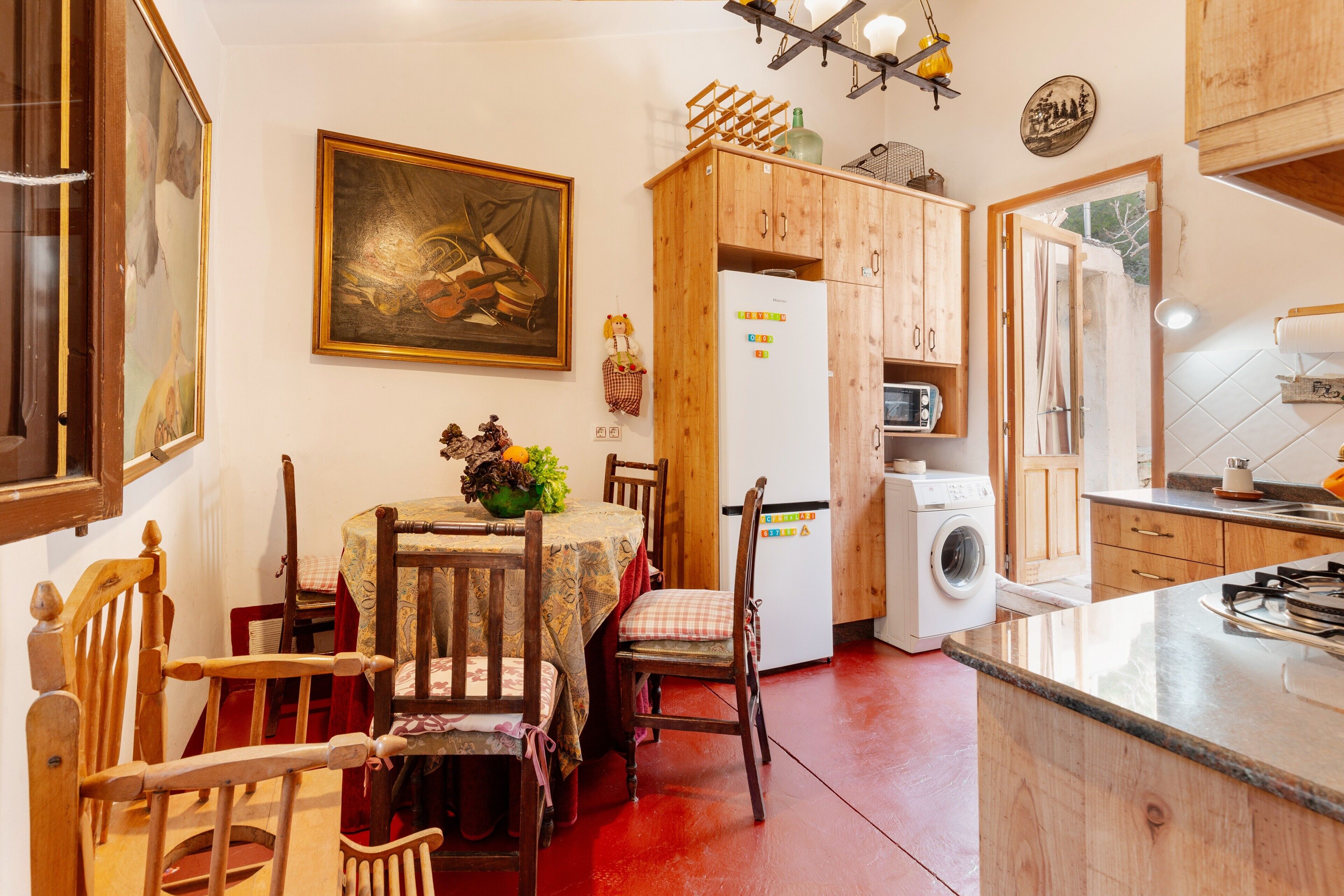 A kitchen with wooden cabinets, a dining table with chairs, a refrigerator, a washing machine, and a painting on the wall.