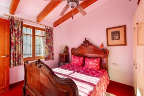 Una camera da letto con un letto in legno, una ventola a soffitto, una finestra con tende floreali e un quadro appeso al muro.