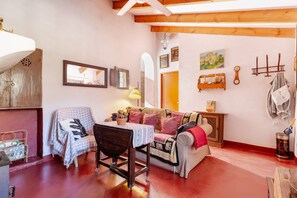 Una stanza accogliente con soffitto in legno, un divano, un tavolo con tovaglia, una sedia, una lampada e una parete decorata con quadri e una mensola con oggetti.