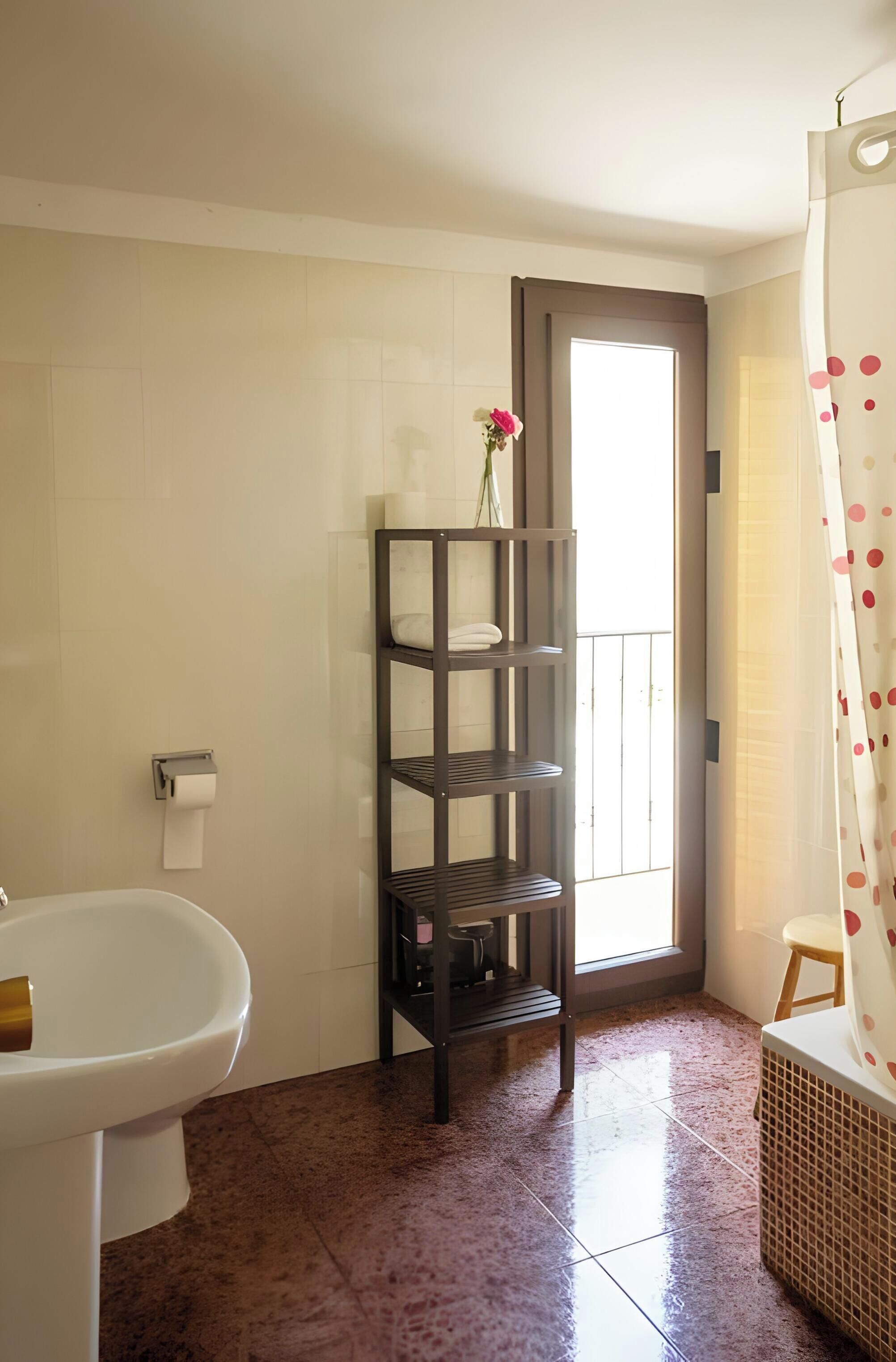 Un baño con un lavabo blanco, un estante de madera oscura con toallas, un dispensador de papel higiénico y una ventana con vistas.