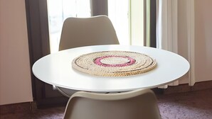 Mesa redonda con un posavasos tejido, dos sillas y vista a un balcón a través de una ventana.