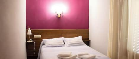 Una cama bien tendida con almohadas y toallas dobladas, una lámpara de noche y una lámpara de pared.