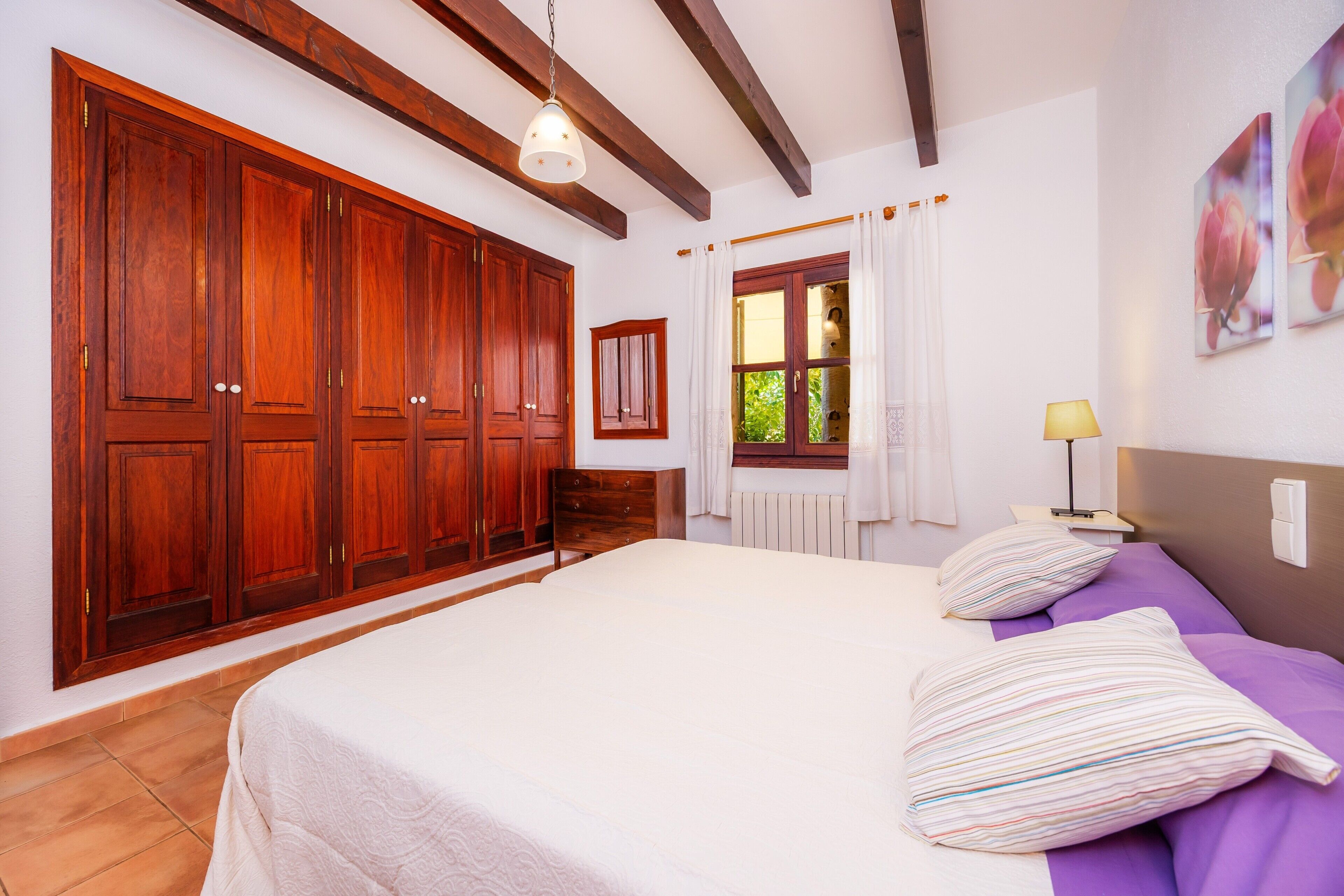 Un dormitorio con un armario grande, una cama con almohadillas a rayas, una mesita de noche con una lámpara y una ventana con cortinas.