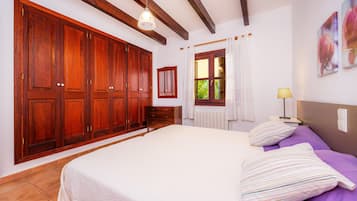 Un dormitorio con un armario grande, una cama con almohadillas a rayas, una mesita de noche con una lámpara y una ventana con cortinas.