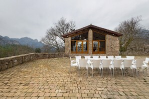 Un bâtiment en pierre avec un toit en bois, de grandes fenêtres en verre et une terrasse dallée avec des chaises blanches.