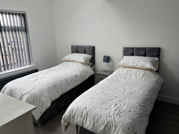 A bedroom with two single beds, a bedside table, a lamp, and a window with blinds.