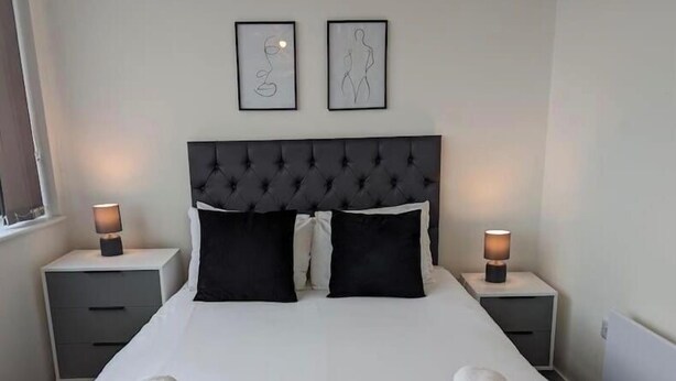 Una cama bien hecha con sábanas blancas y almohadas negras, dos mesitas de noche con lámparas, y dos cuadros en la pared.
