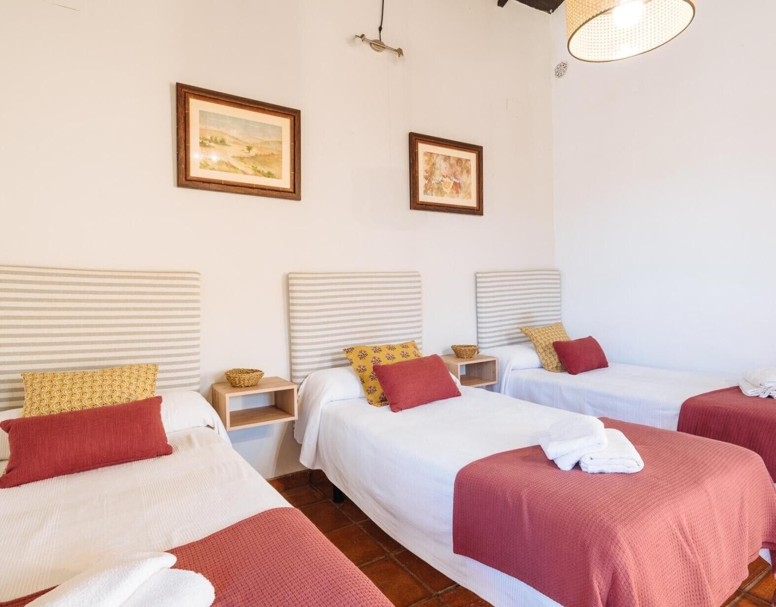 A hotel room with three beds, each with a red blanket, white bedding, and a small bedside table with a basket. There are two framed pictures on the wall.