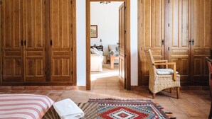 A room with wooden wardrobes, a wicker chair, a patterned rug, and a bed with a striped bedspread. - Finca Los Gorriones – Country House with Mountain Views near Seville (El Garrobo)
